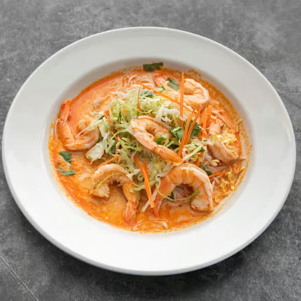 A white bowl of Mai Thai shrimp laksa with noodles, shredded cabbage, and carrots in a red curry broth.